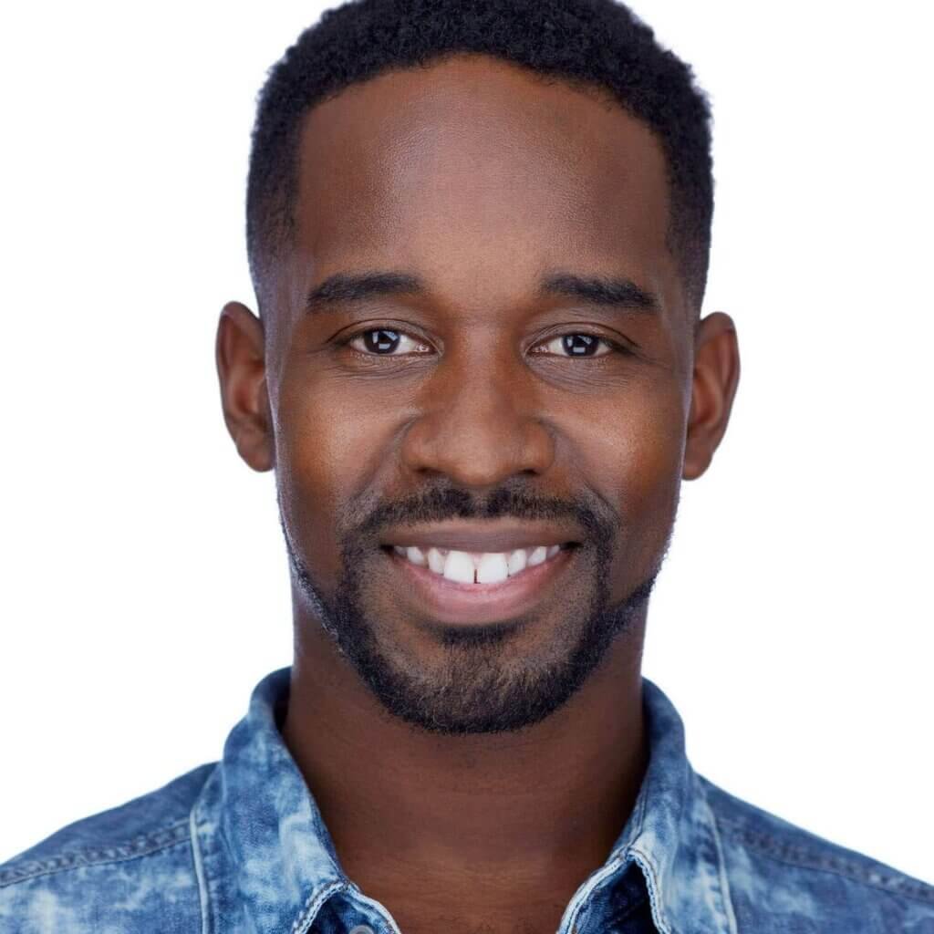 A confident man smiles at the camera against a white background. He has short black hair, a beard, and wears a blue denim shirt. His friendly, approachable expression suggests he might be an actor and the headshot was taken in Stamford, CT.