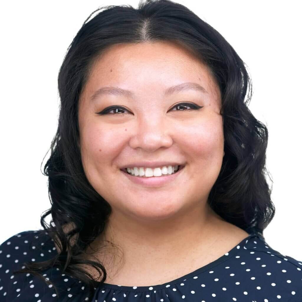 Smiling Asian woman with long, wavy black hair against a white background. Her headshot features a navy blue blouse adorned with white polka dots and subtle, winged eyeliner.