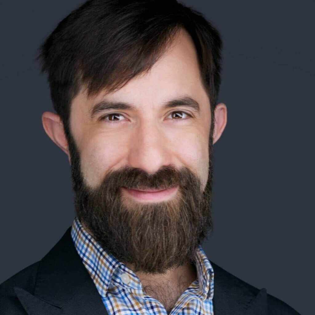 A man with a full beard and mustache is smiling softly in this professional headshot. He wears a dark suit jacket over a blue and white checkered shirt. His dark hair is neatly styled against the smooth, solid dark gray background, accentuating his features.