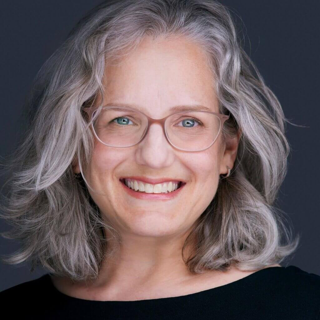 A person with shoulder-length gray hair and glasses exudes confidence as they smile warmly against a dark background. Their black top complements their light blue eyes, while the lighting enhances this engaging headshot, capturing a friendly expression.