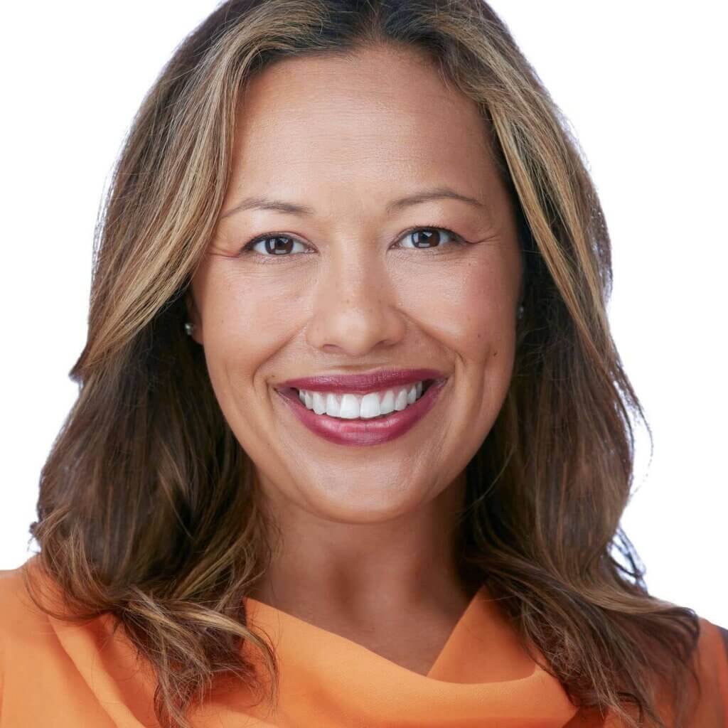 Smiling female executive with shoulder-length brown hair, wearing an orange top. The plain white background highlights her cheerful expression and casual yet professional attire in this engaging headshot.