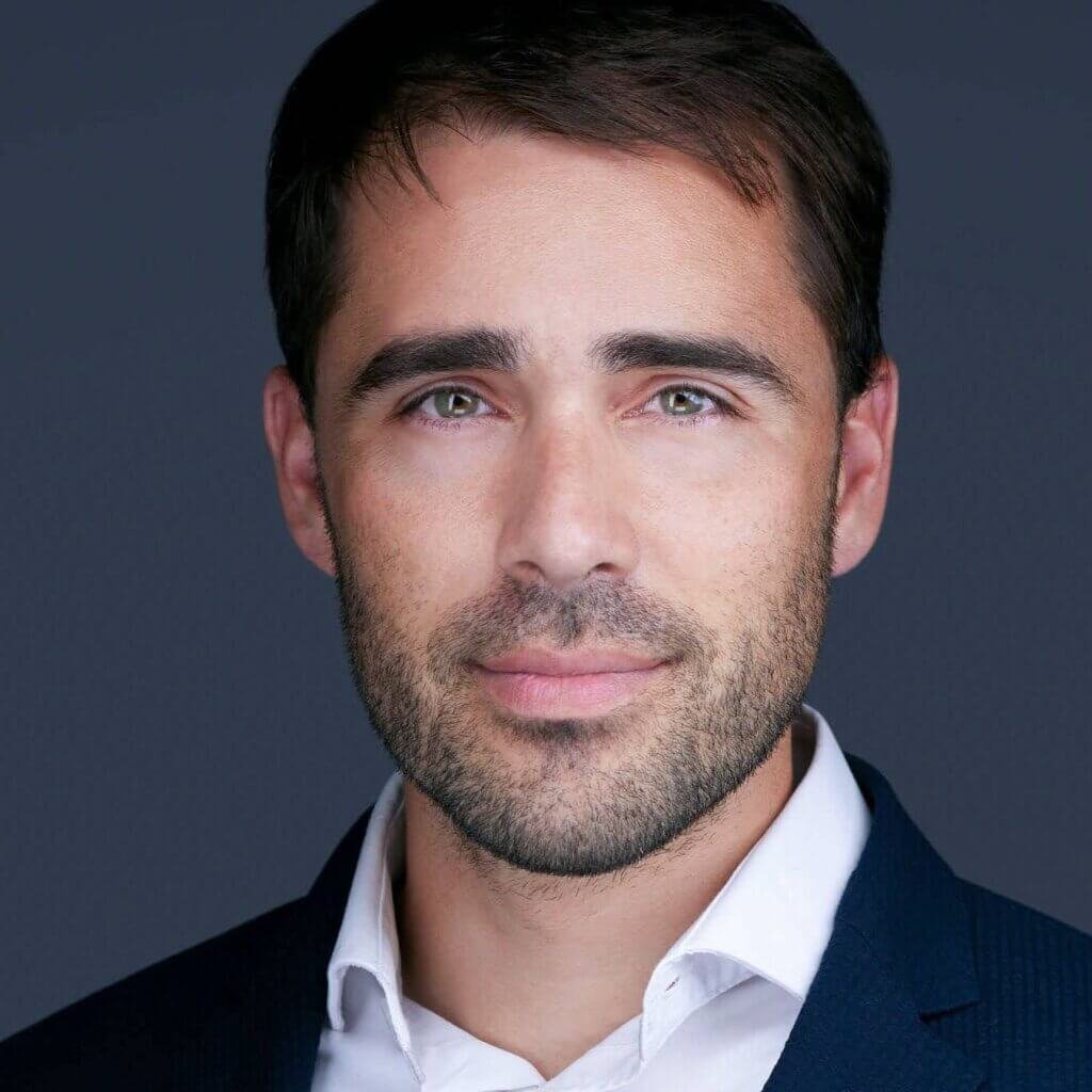 A consultant's headshot captures a man with short, dark hair and a beard, wearing a navy suit and white shirt. His serious expression and calm demeanor are emphasized by his direct eye contact. The neutral gray backdrop keeps the focus on the subject's face.