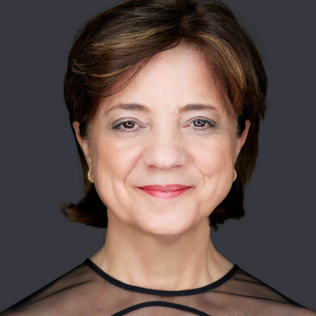 A woman with short brown hair smiles softly in her headshot against a dark gray background. She is wearing a sheer black top and gold earrings, subtly enhanced by pink lipstick and light eye makeup. The lighting highlights her features, crafting a warm, approachable image suitable for the board of directors.