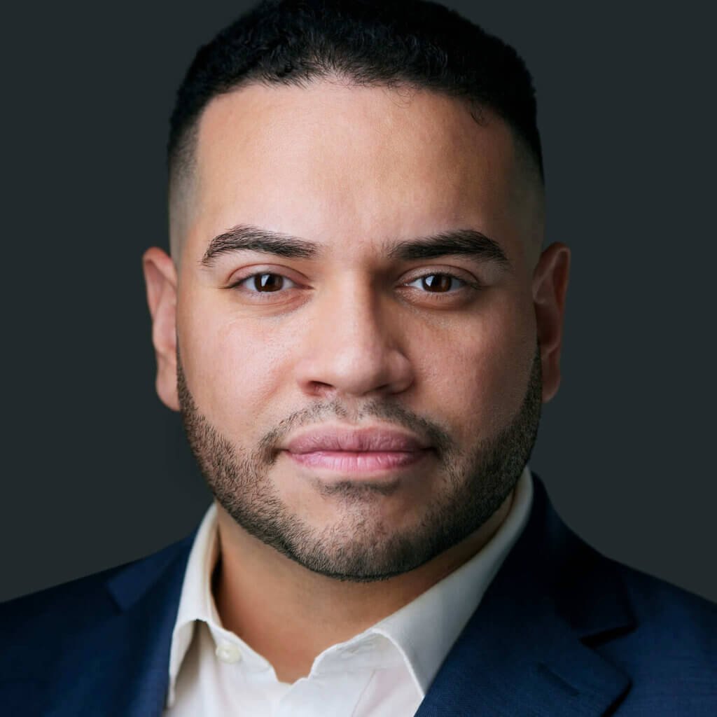 A professional headshot of an approachable, confident consultant with short black hair and a trimmed beard, wearing a dark blue suit and white shirt against a grey background.