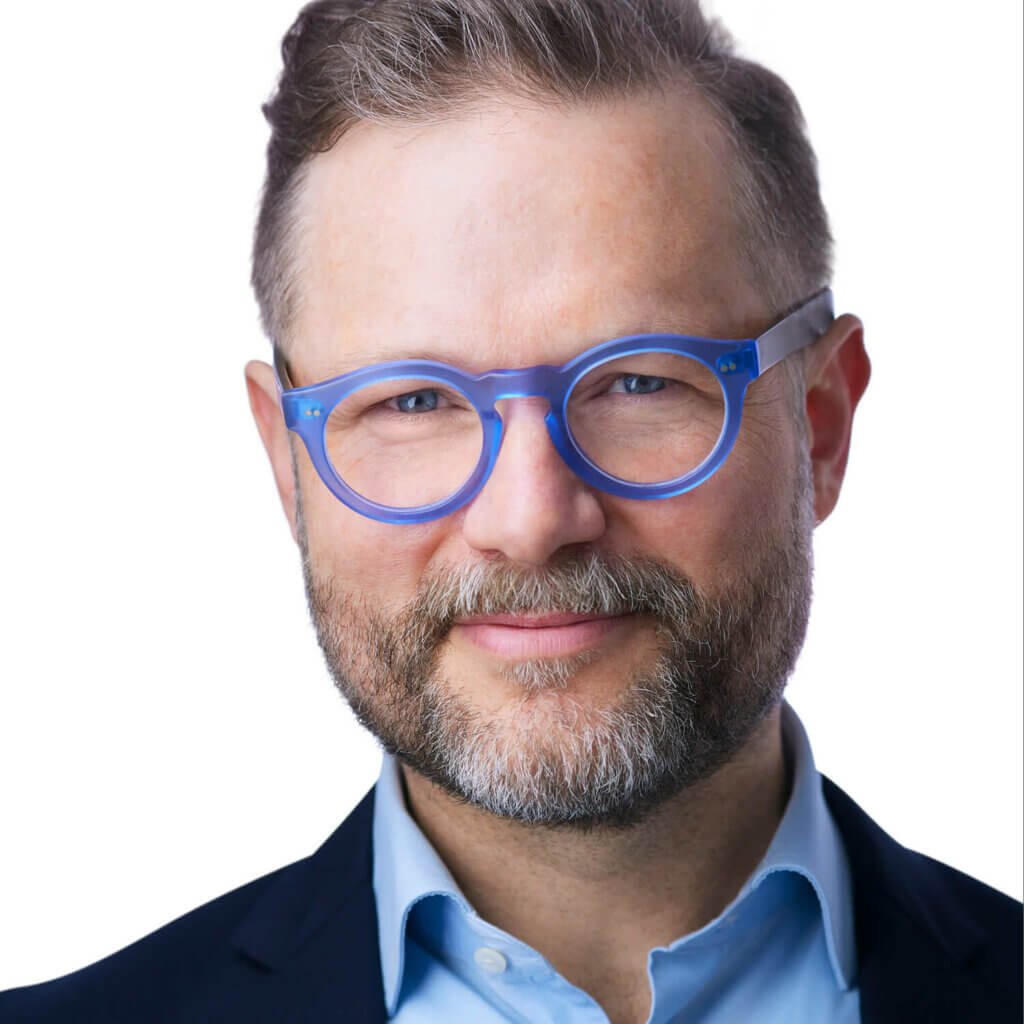 A middle-aged man, resembling a CMO, with neatly groomed short hair and a beard, wears round blue glasses and a light blue collared shirt under a dark blazer. His headshot captures a slight smile against a plain white background.