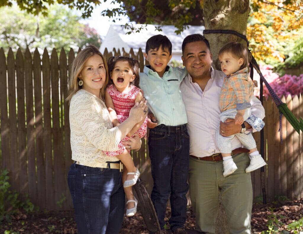 Photographer Hector Pachas and family. His family of five stands outdoors near a wooden fence and tree. The mother is holding a young girl in a red dress, both smiling. The father, wearing a beige shirt, holds a toddler in a plaid shirt. A young boy in a light blue shirt stands between the parents, and smiles with one arm on his dad.