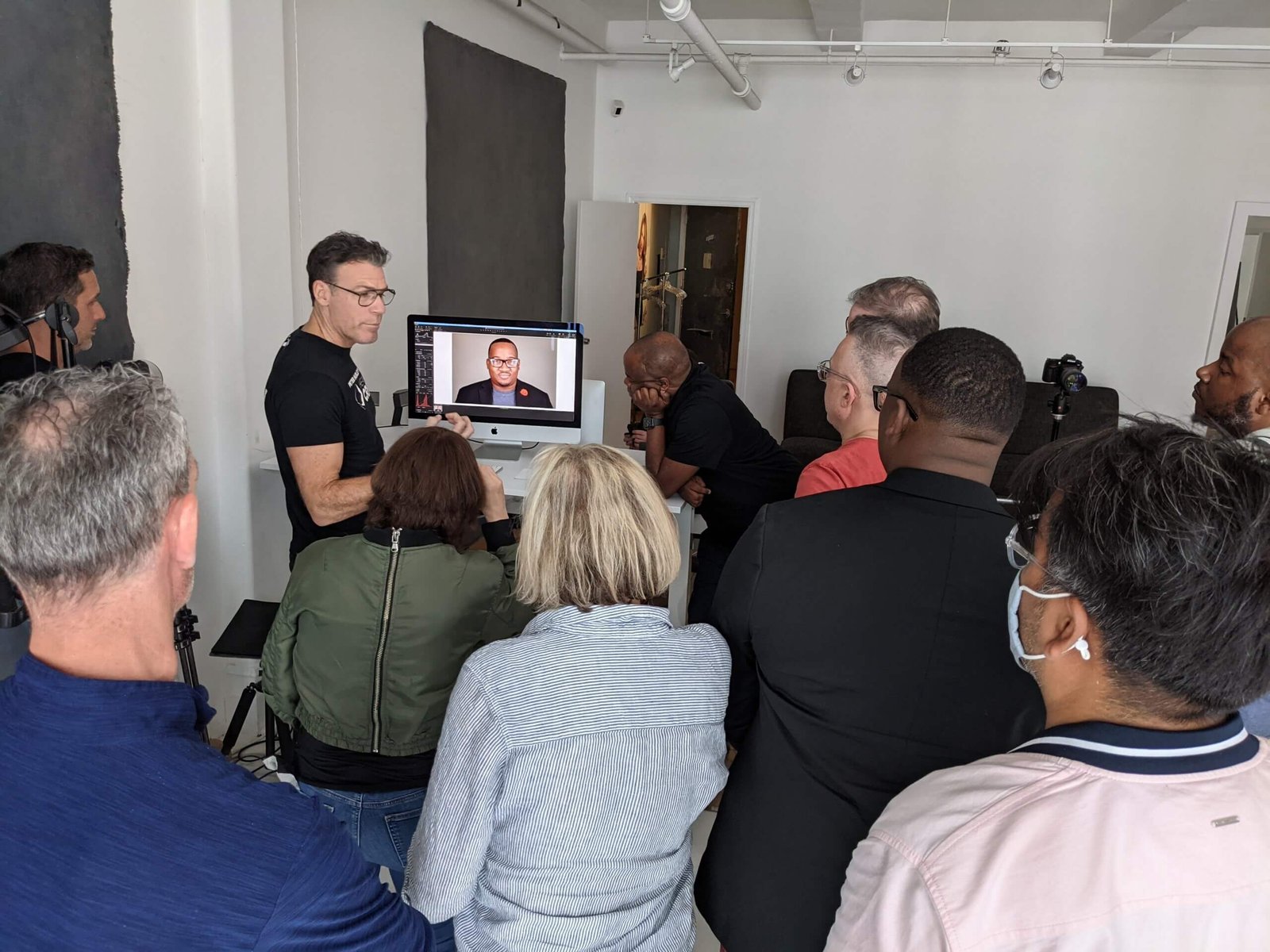 A group of people is gathered in a room, attentively watching a person present something on a computer screen. The presenter, standing near a desk, is showing an image of a male face on the monitor. The attendees are standing close together, some wearing glasses and one person wearing a mask.