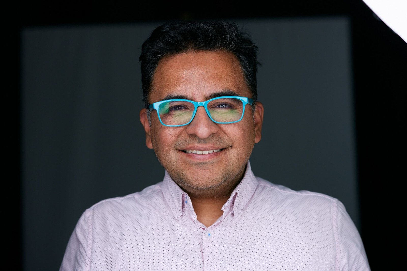 A man with short black hair, wearing bright blue glasses and a light pink button-down shirt, is smiling at the camera for a modern headshot. The background is dark, likely a studio setting, and he's illuminated by soft lighting that highlights his face.